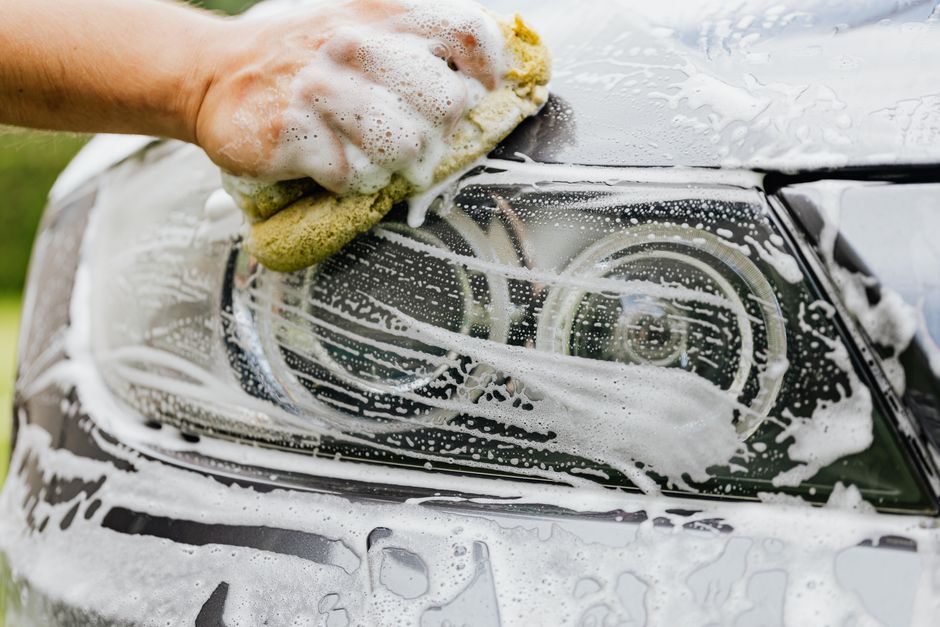 Hand scrubbing a car headlight with sponge and soap suds, detailing process.