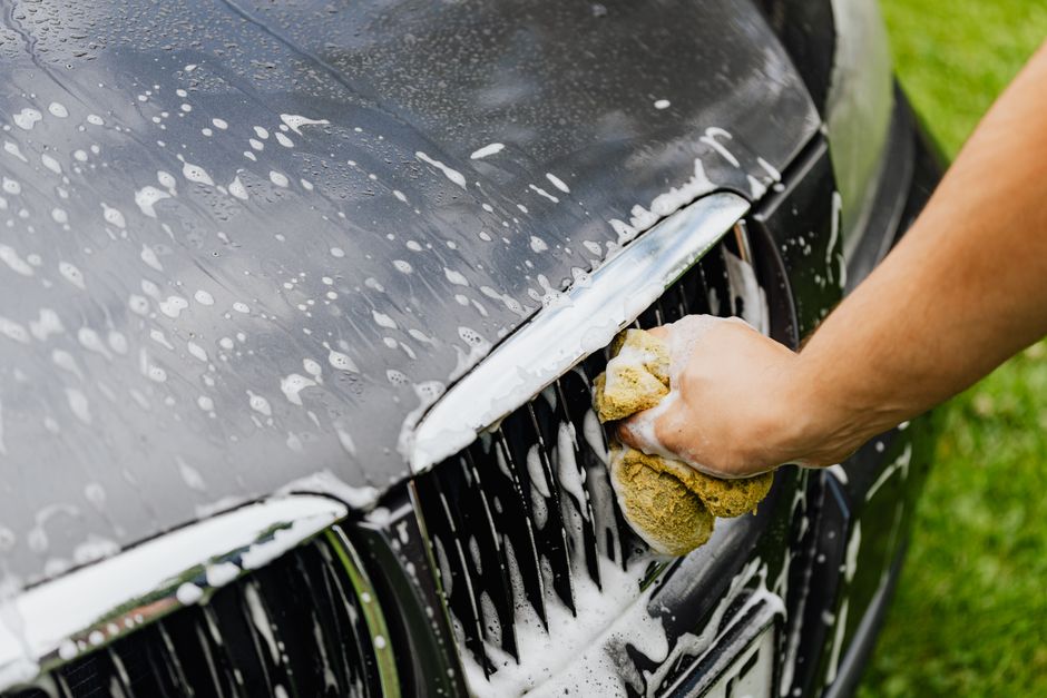 Detailed view of a hand washing a car grille with a soapy sponge outdoors.