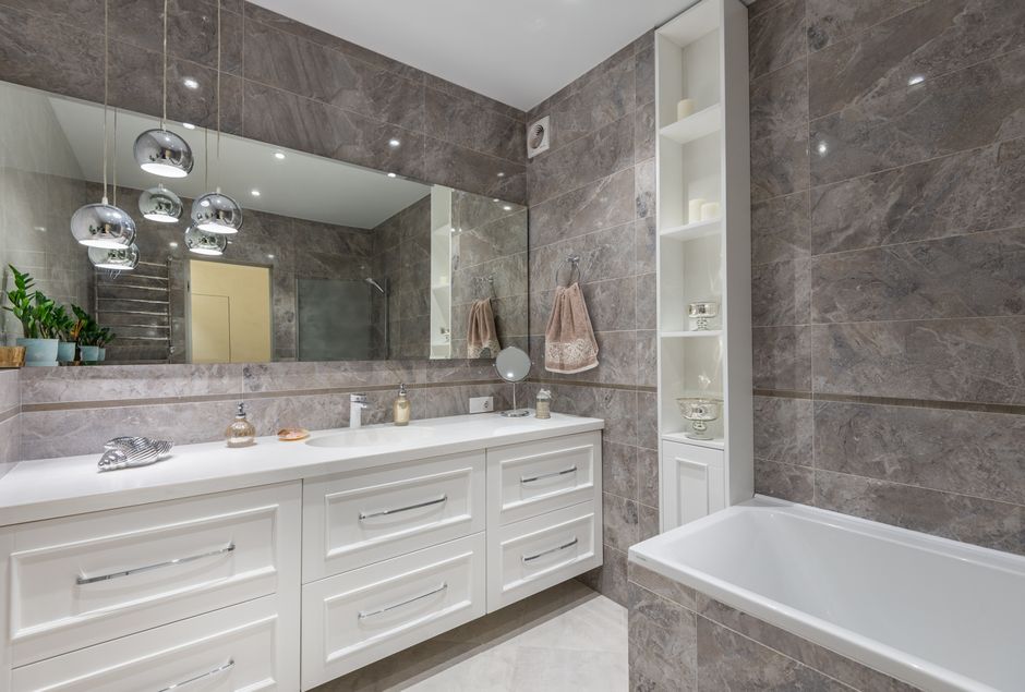 Big window hanging on tiled wall above while cabinet and sink near bathtub in modern bathroom decorated with creative lamps and potted plants