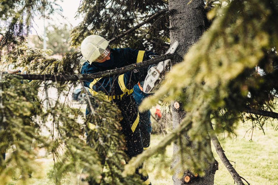 Firefighter in protective gear using chainsaw to cut tree branches outdoors.