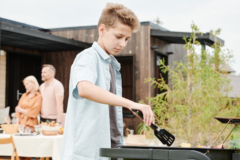 A young boy grills food during a family barbecue in the backyard, with adults in the background.