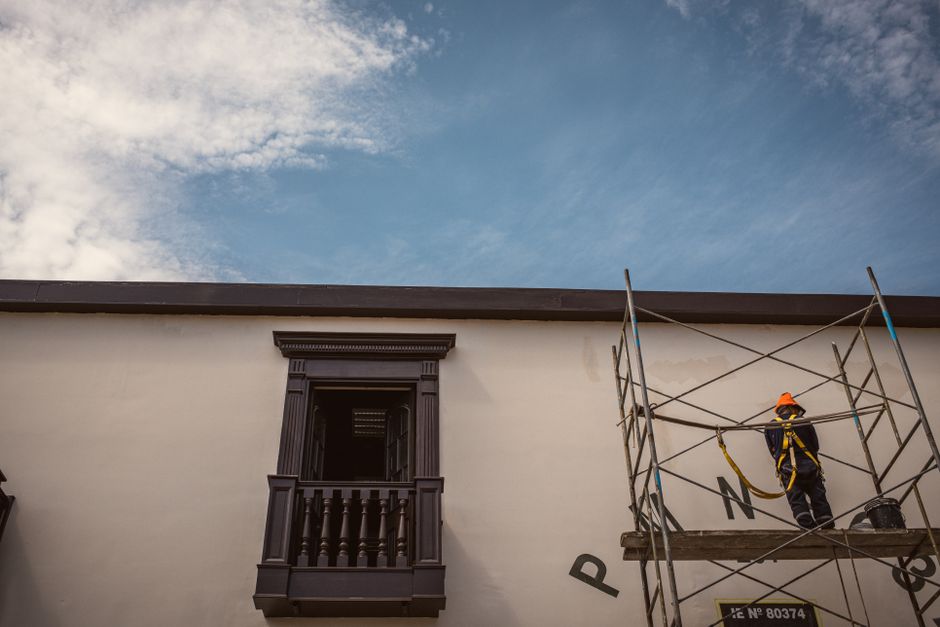 A construction worker wearing safety gear stands on scaffolding beside a traditional building.