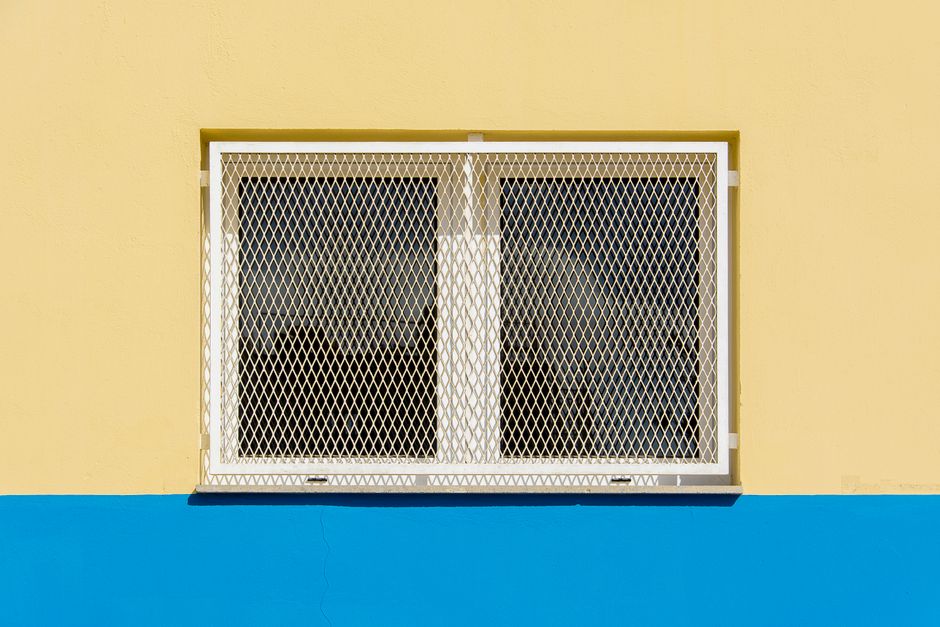 A yellow building facade featuring a barred window above a blue stripe, offering contrast and texture.