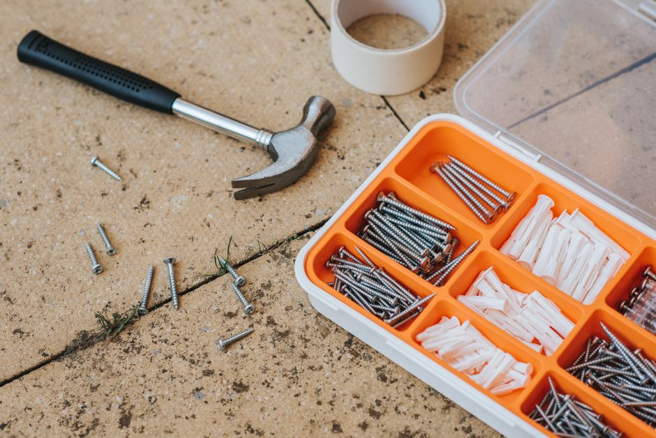 Top view of assorted repair instruments and screws in box near hammer and scotch tape on dirty tile in soft daylight