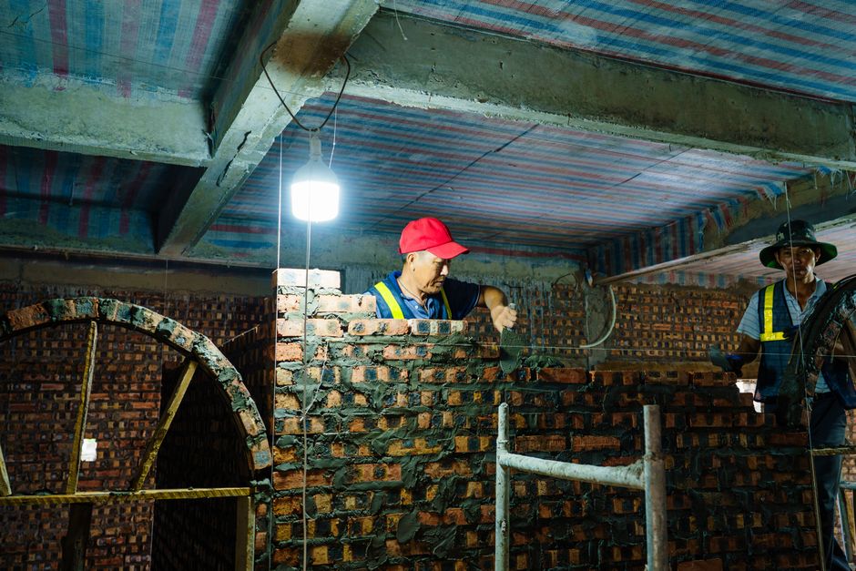 Workers constructing a brick wall indoors under a dimly lit environment in Hải Phòng, Việt Nam.