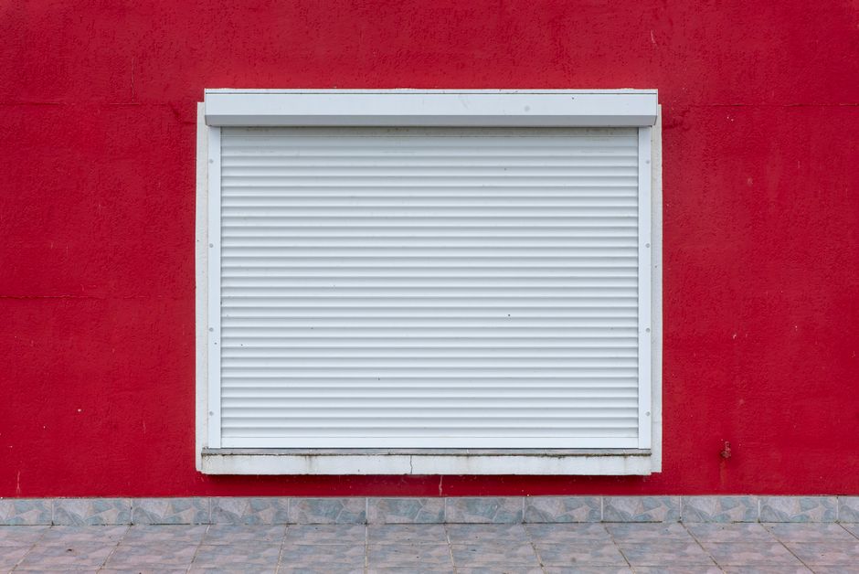 A minimalist exterior view of a white roller shutter on a vibrant red wall.