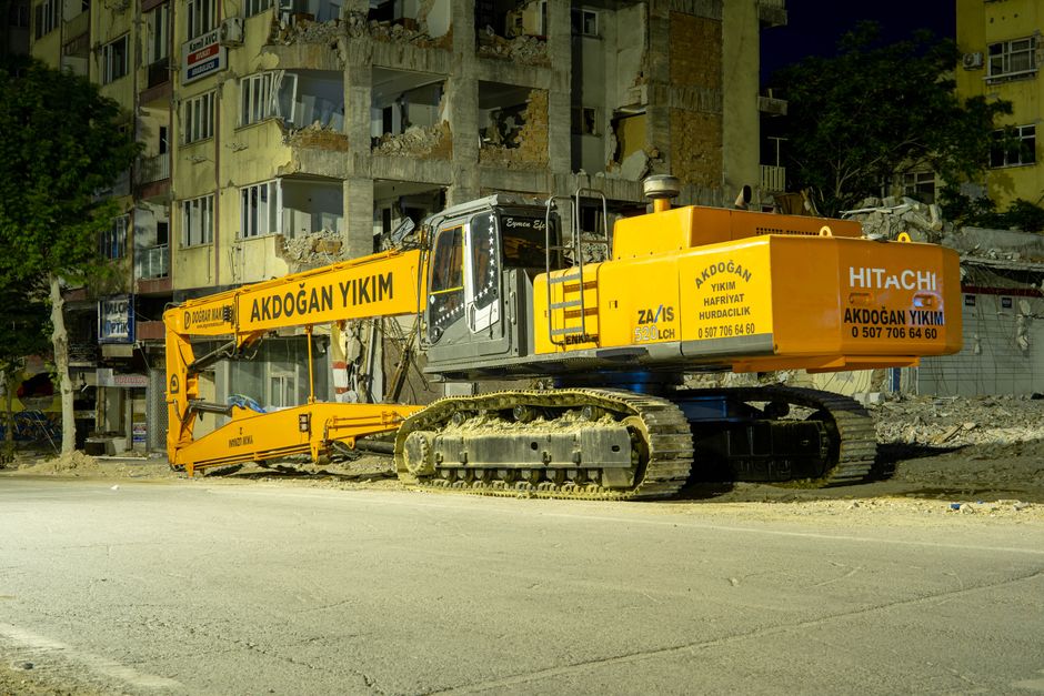 Large excavator at a city demolition site during nighttime, illuminated by streetlights.
