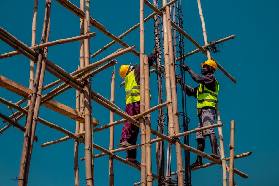 Two construction workers on bamboo scaffolding, wearing safety gear under a clear blue sky.
