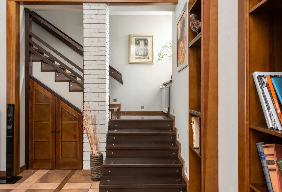 Wooden stairway near brick column placed at shelves with books in modern flat with pictures on wall and dry plants in vase