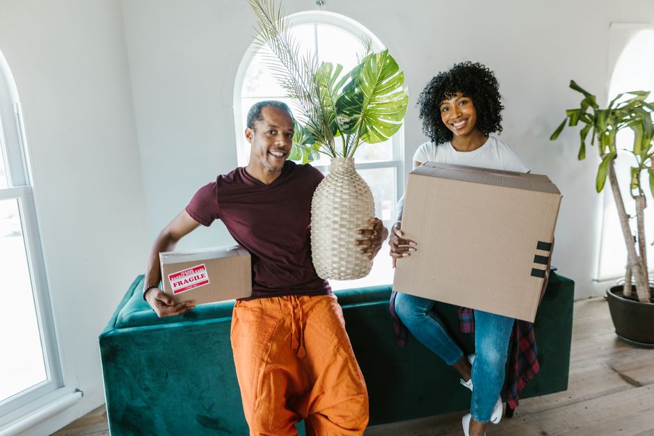 Couple smiling while moving into a new home, carrying boxes and a plant.