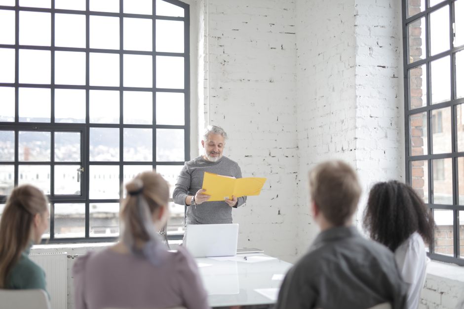 Aged gray haired bearded male mentor in casual outfit presenting ideas at multiracial group meeting in creative office workspace while standing at table with laptop